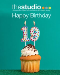 A cupcake with white icing and chocolate sprinkles has two lit birthday candles shaped as the number 19. Above, text reads thestudio Happy Birthday on a green background.