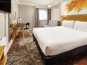 Modern hotel room with a large bed, desk, chair, sofa, wall art, and a window with grey curtains. The carpet has a geometric pattern with orange and grey colours.