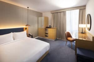 Modern hotel room with a neatly made double bed, wooden furniture, desk with chair, wardrobe, coffee station, large window with curtains, and soft lighting.