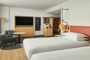 Modern hotel room with two white beds, a green armchair, round table, flat-screen TV, and kitchenette with microwave, mini fridge, and storage. Walls are white with orange accents. Room is bright with natural light.