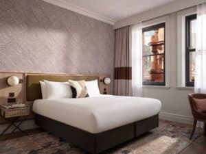 A modern hotel room with a double bed, geometric feature wall, two windows, a bedside table with a glass of water, curtains, and an upholstered chair on a patterned carpet.