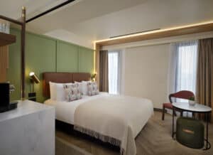 Modern hotel room with a large bed, colourful pillows, green accent wall, small round table with two chairs, floor-to-ceiling windows with sheer curtains, and wooden flooring. Bright and inviting atmosphere.