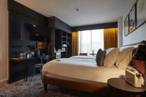 Modern hotel room with a large bed, bedside table, TV on a dark wall, desk with chair, microwave, and large window with yellow curtains letting in natural light.