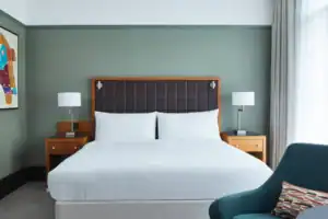 Modern hotel room with a large bed, white striped bedlinen, two wooden bedside tables with lamps, a teal chair, and a window with beige curtains. A colourful abstract painting hangs on the wall.